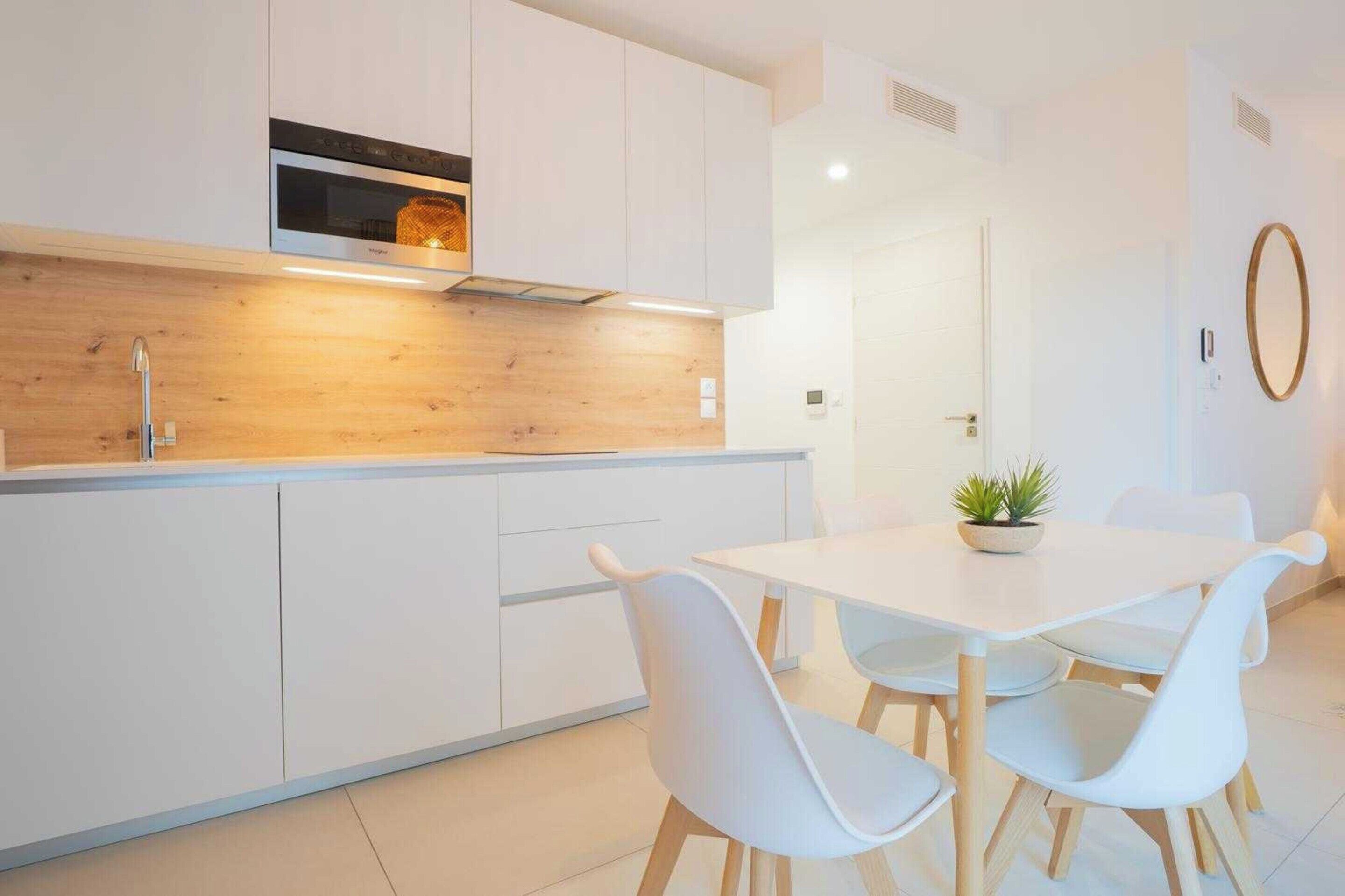 Una cocina moderna con armarios blancos, un protector de salpicadero de madera y una zona de comedor con mesa y sillas blancas.