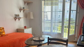 Een kamer met een bed, een tafel, een stoel, een lamp, een raam met gordijnen en uitzicht op gebouwen.