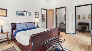 A bedroom with a wooden bed, a nightstand, a lamp, a mirror, and a piano in the adjacent room. - Riverside Retreat! Quiet and Charming 2 bed/2 bath (New Orleans)