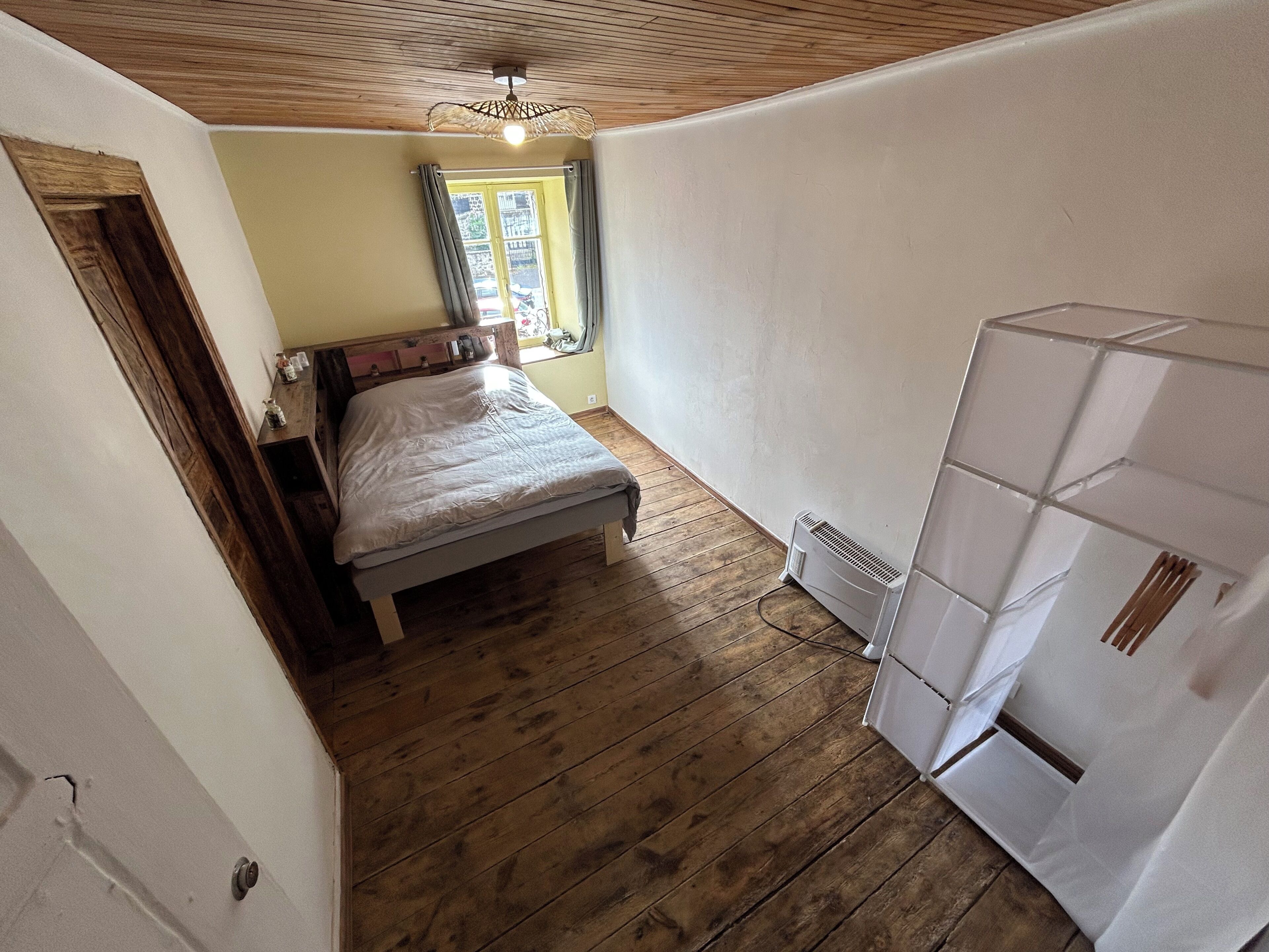 A bedroom with a wooden floor, a bed, a window with curtains, a ceiling fan, and a white radiator.