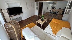 A living room with a sofa, a television, a small table with a plant, and a kitchen area in the background. - Room in a house 5 minutes from Lac du Bouchet (Cayres)