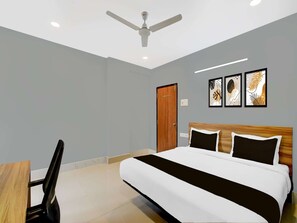 A hotel room with a bed, a desk, a chair, a ceiling fan, and artwork on the wall. - Deluxehotel Santosh Palace (Prayagraj)