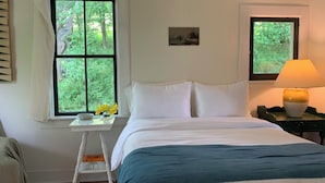 Un dormitorio con cama, ventana con vista a follaje, mesita de noche con lámpara y una pequeña mesa con un florero.