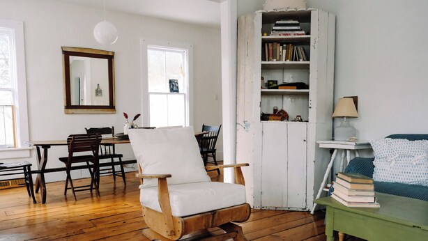 Una habitación con una mecedora de madera, una mesita auxiliar verde con libros, un armario blanco con libros y una ventana con vistas.