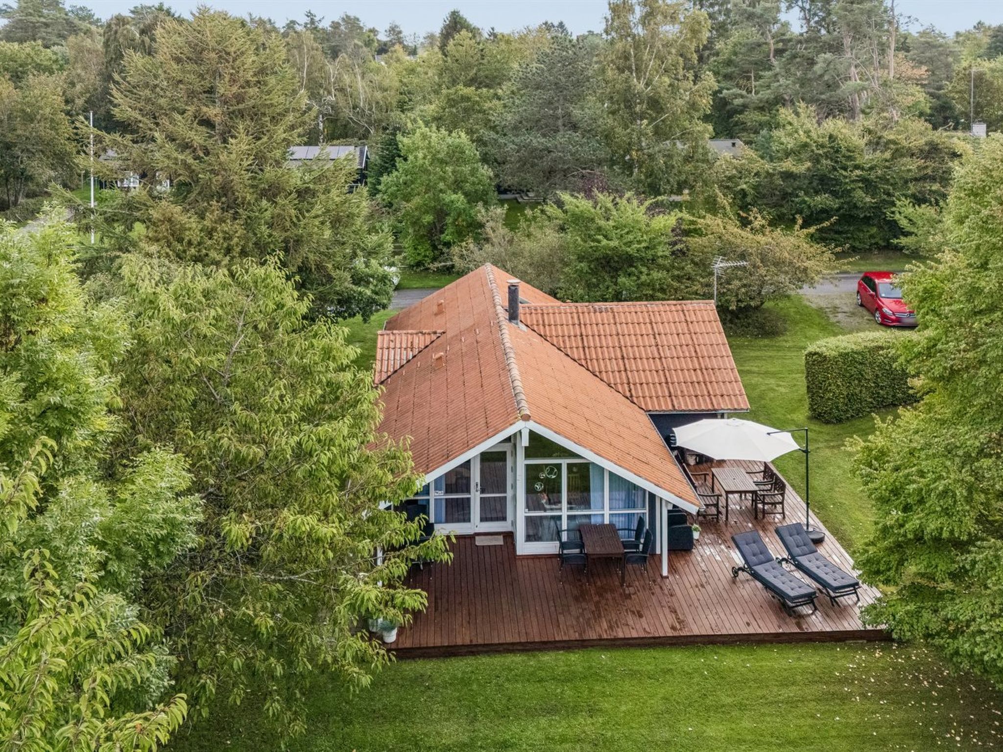 A house with a sloped roof, a deck with outdoor furniture, and a red car parked nearby.