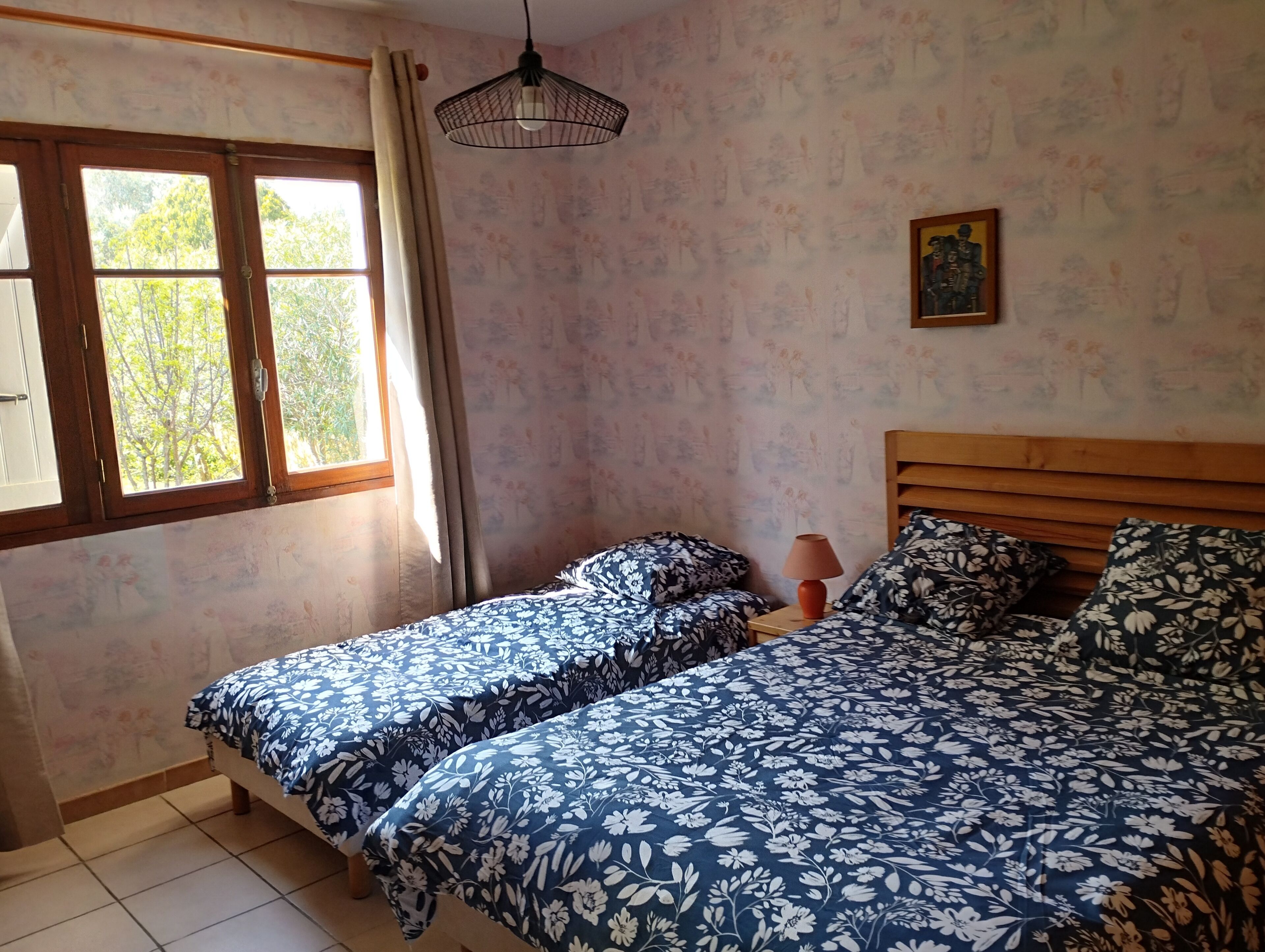 Una stanza con due letti, una finestra con tende, una lampada a sospensione e un quadro appeso al muro.