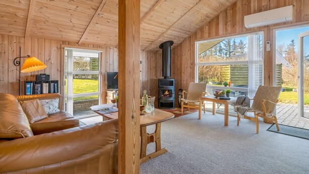 A cozy cabin interior with a fireplace, leather sofa, wooden tables, and large windows.
