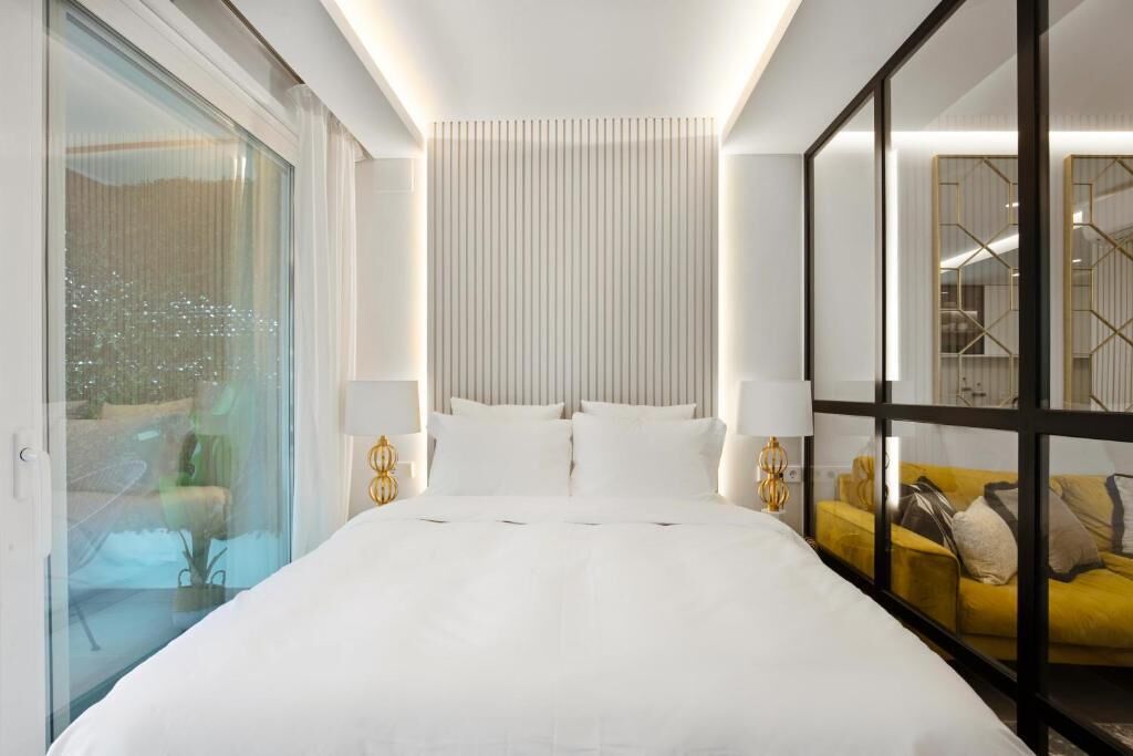 Camera d'albergo con un letto grande, due comodini con lampade, un divisorio in vetro e un divano giallo.