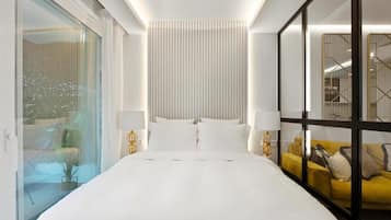 Habitación de hotel con una cama grande, dos mesitas de noche con lámparas, un separador de vidrio y un sofá amarillo.