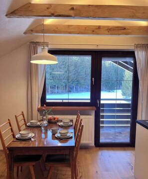 Een eetgedeelte met een houten tafel, stoelen, een hanglamp en uitzicht op bomen door een groot raam.
