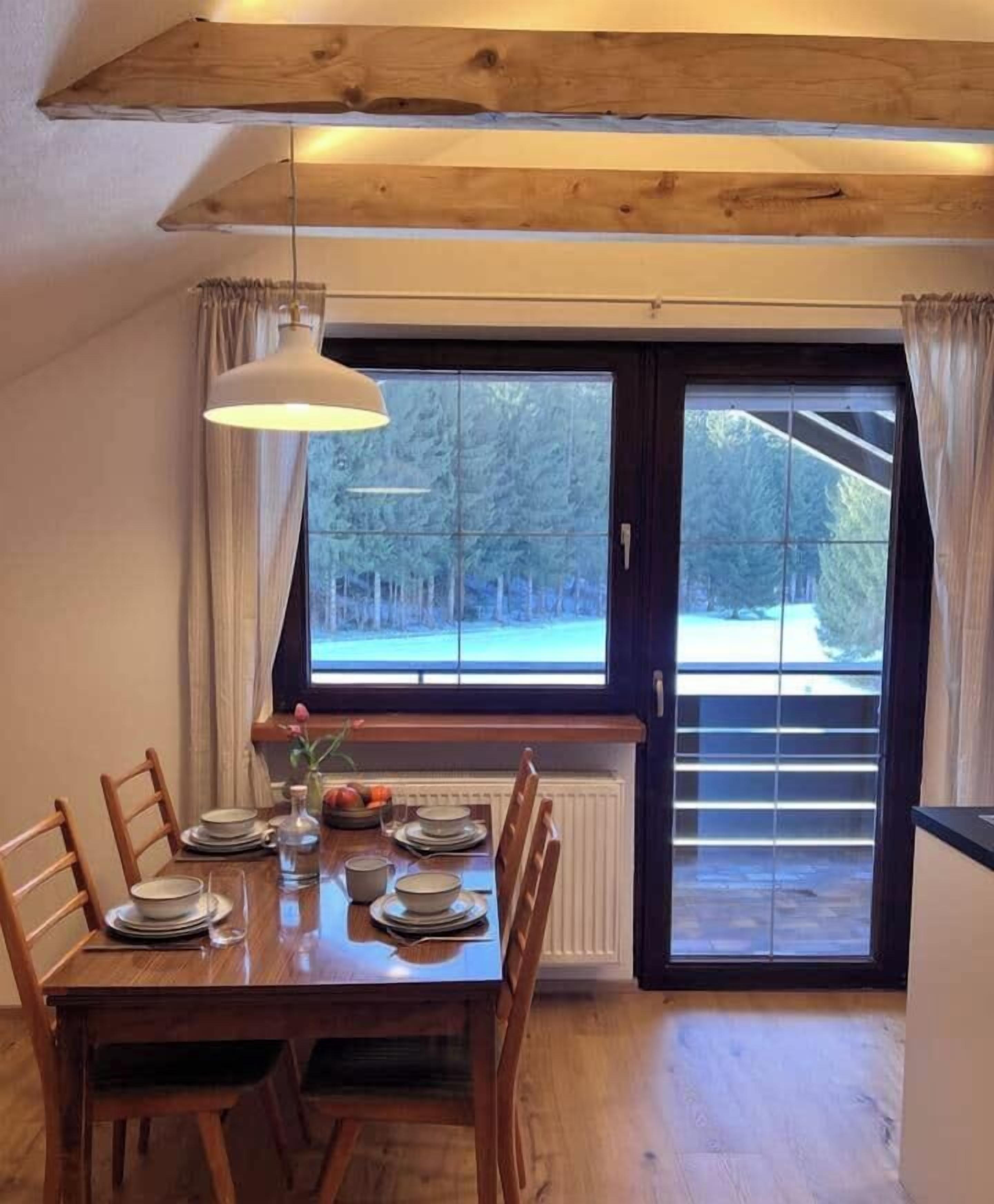 Ein Essbereich mit einem Holztisch, gedeckt für vier Personen, einer hängenden Pendelleuchte und Blick durch ein großes Fenster.