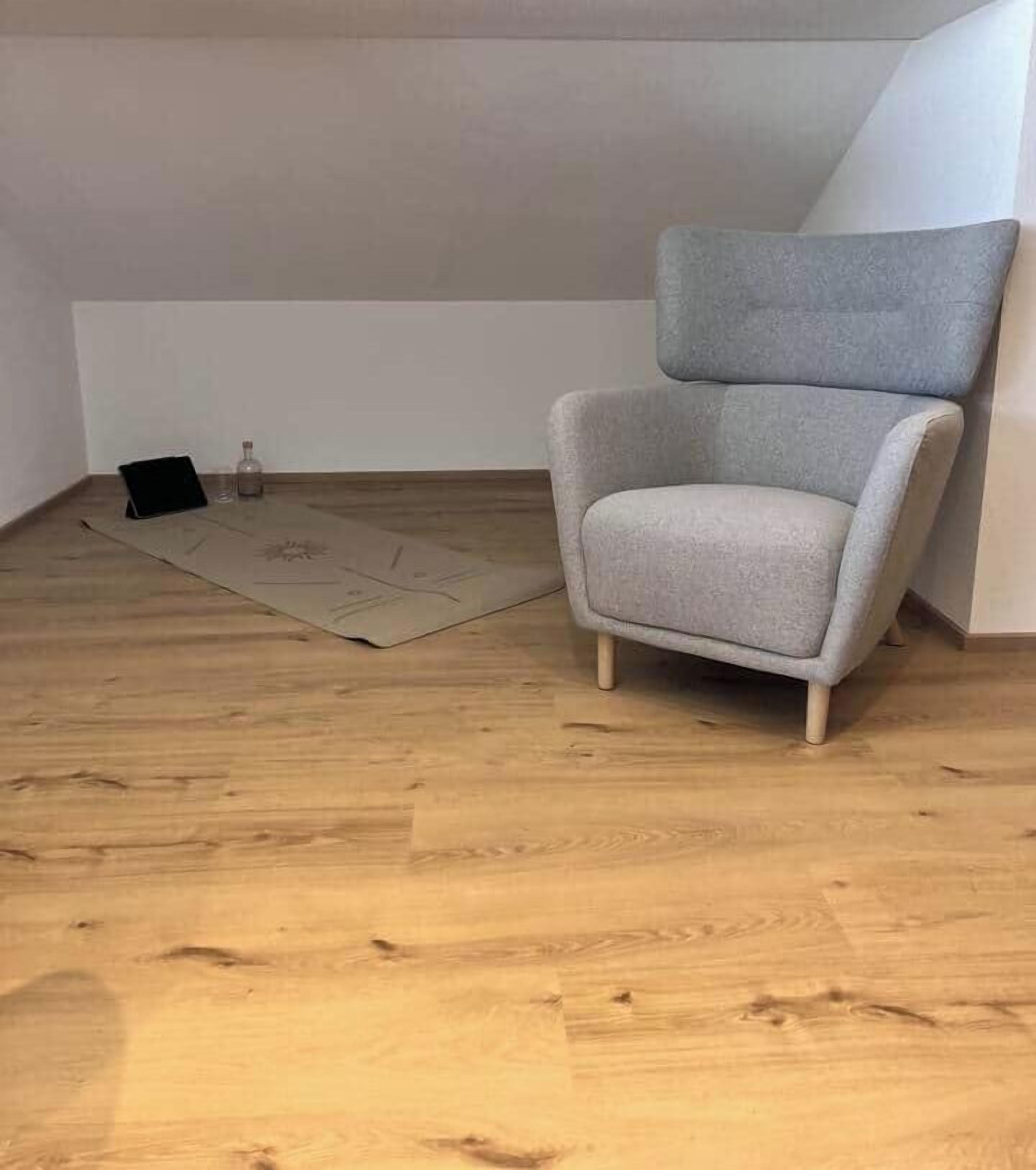 Ein Zimmer mit einem grau gepolsterten Sessel, Holzboden und einem kleinen Tisch mit einer Flasche und einem schwarzen Gegenstand.