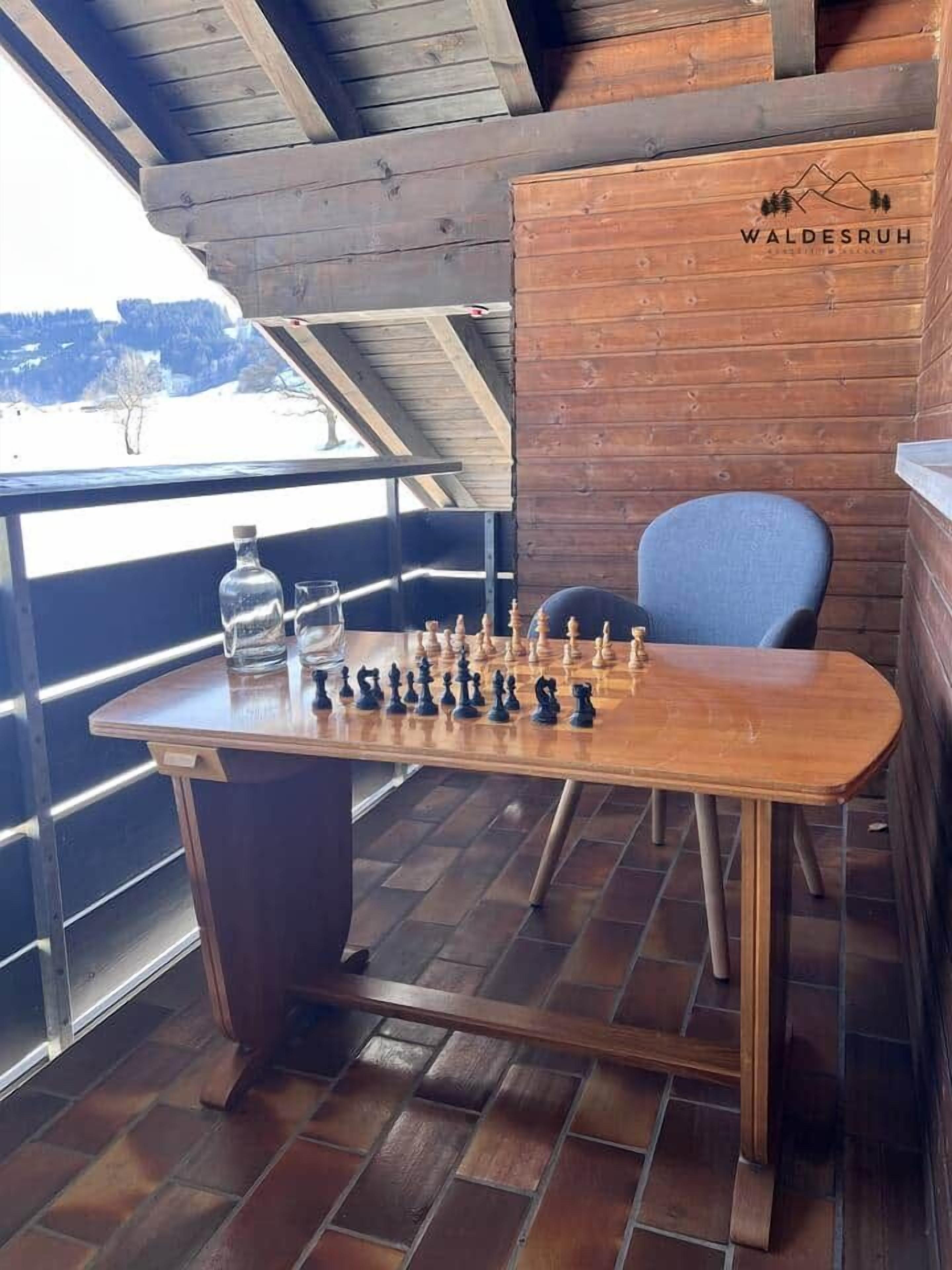 A wooden table with a chessboard, a chair, and a bottle on a balcony with a view.