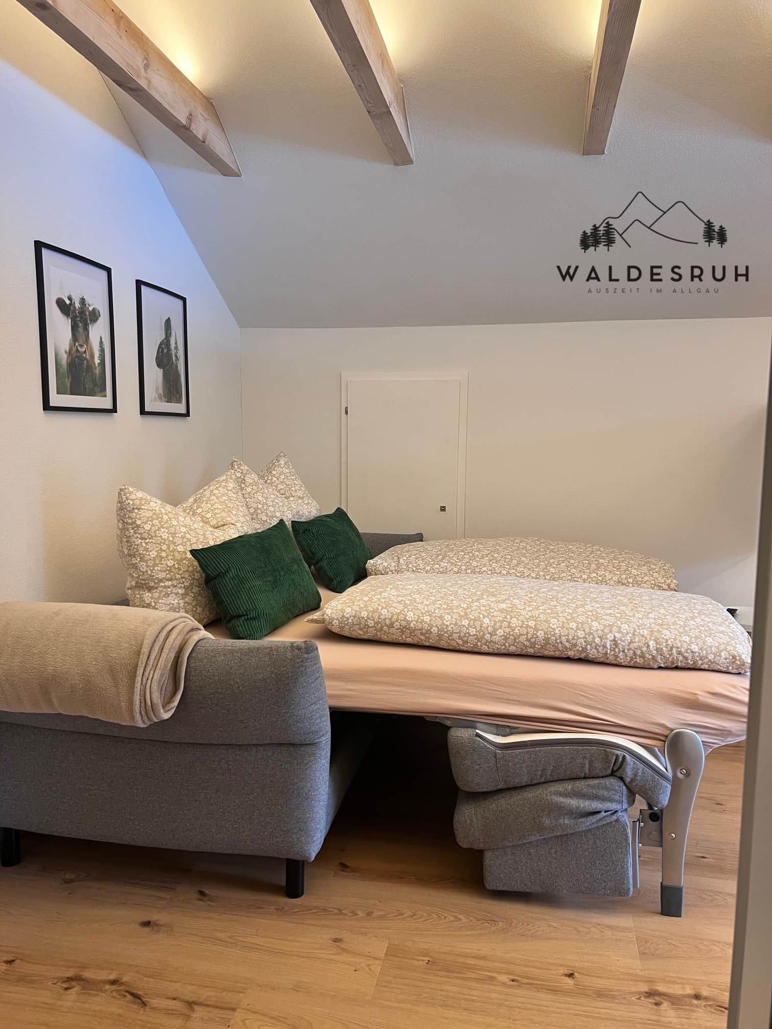 A modern bedroom with a bed, a sofa, and a wheelchair. There are framed pictures on the wall and a logo on the ceiling.
