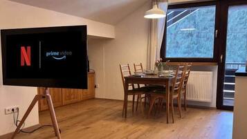 A modern living room with a flat-screen TV displaying the Netflix logo, a dining area with wooden furniture, and a large window offering a view of trees.
