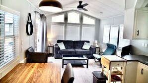 Ett modernt vardagsrum med en soffa, ett soffbord och ett matbord. Det finns en takfläkt, stora fönster med persienner och en köksdel med skåp.
