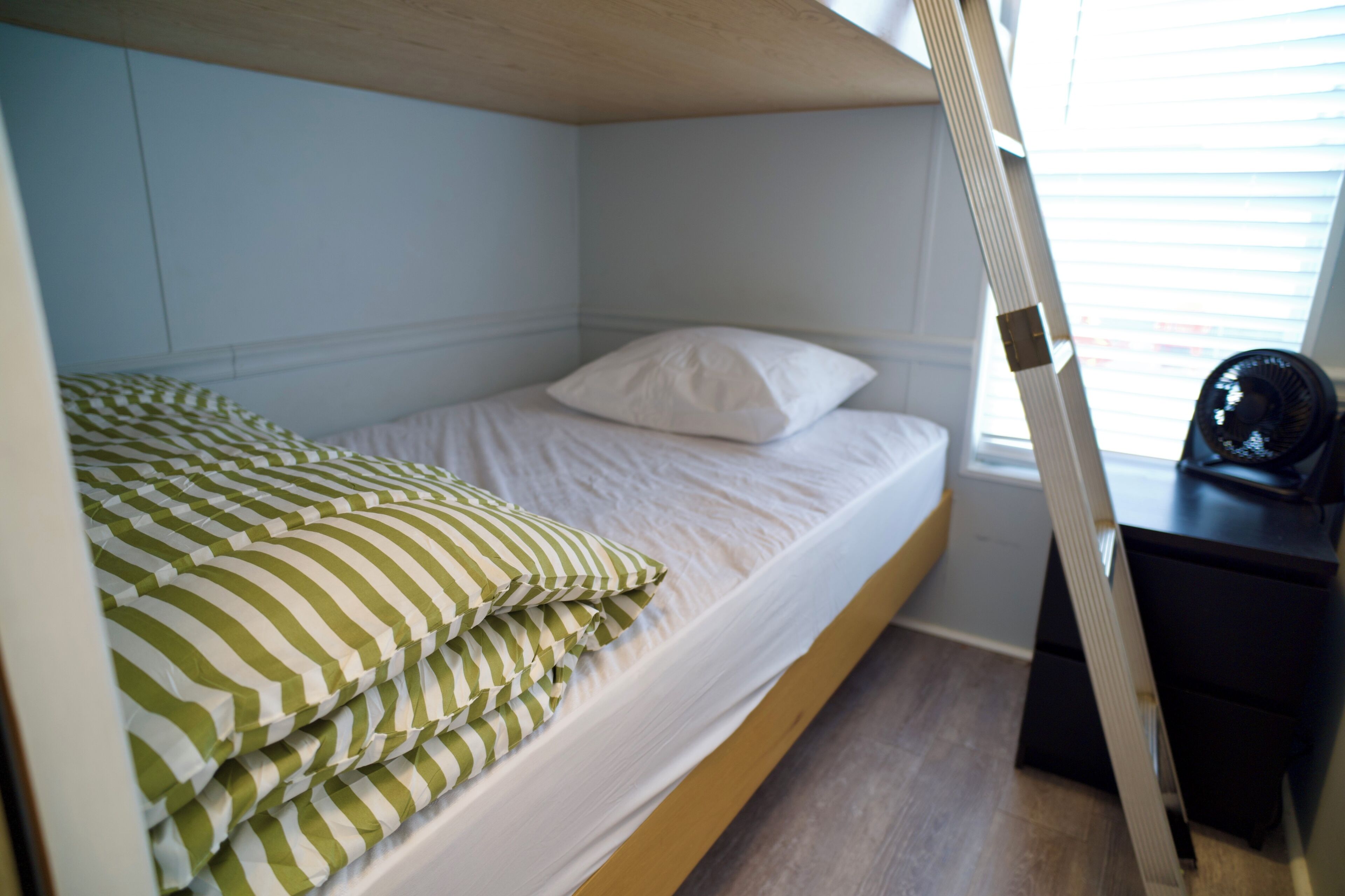 A bunk bed with a white mattress and green striped pillows, a wooden headboard, and a desk with a chair.