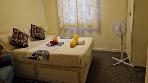 A bedroom with a bed, pillows, a fan, a window with curtains, and a door. - Basildon Holiday Home Essex UK (Basildon)