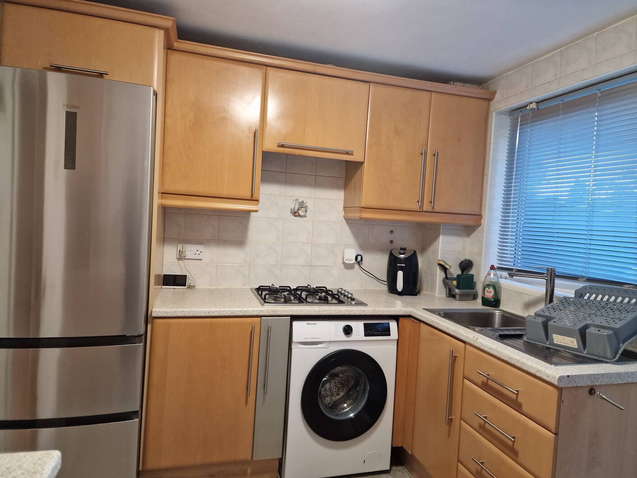 A compact kitchen with wooden cabinets, a stainless steel refrigerator, a washing machine, a gas stove, and a window with blinds.