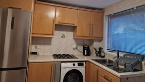 A compact kitchen with wooden cabinets, a stainless steel refrigerator, a washing machine, a gas stove, and a window with blinds. - Basildon Holiday Home Essex UK (Basildon)