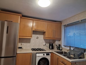 A compact kitchen with wooden cabinets, a stainless steel refrigerator, a washing machine, a gas stove, and a window with blinds.