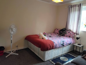 Een slaapkamer met een bed, een ventilator, een tafeltje en een raam met gordijnen.