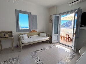 Ein Zimmer mit einer Couch, einem kleinen Tisch, einem Fenster mit Rollläden und einer Tür zu einem Außenbereich mit Tisch und Stühlen.