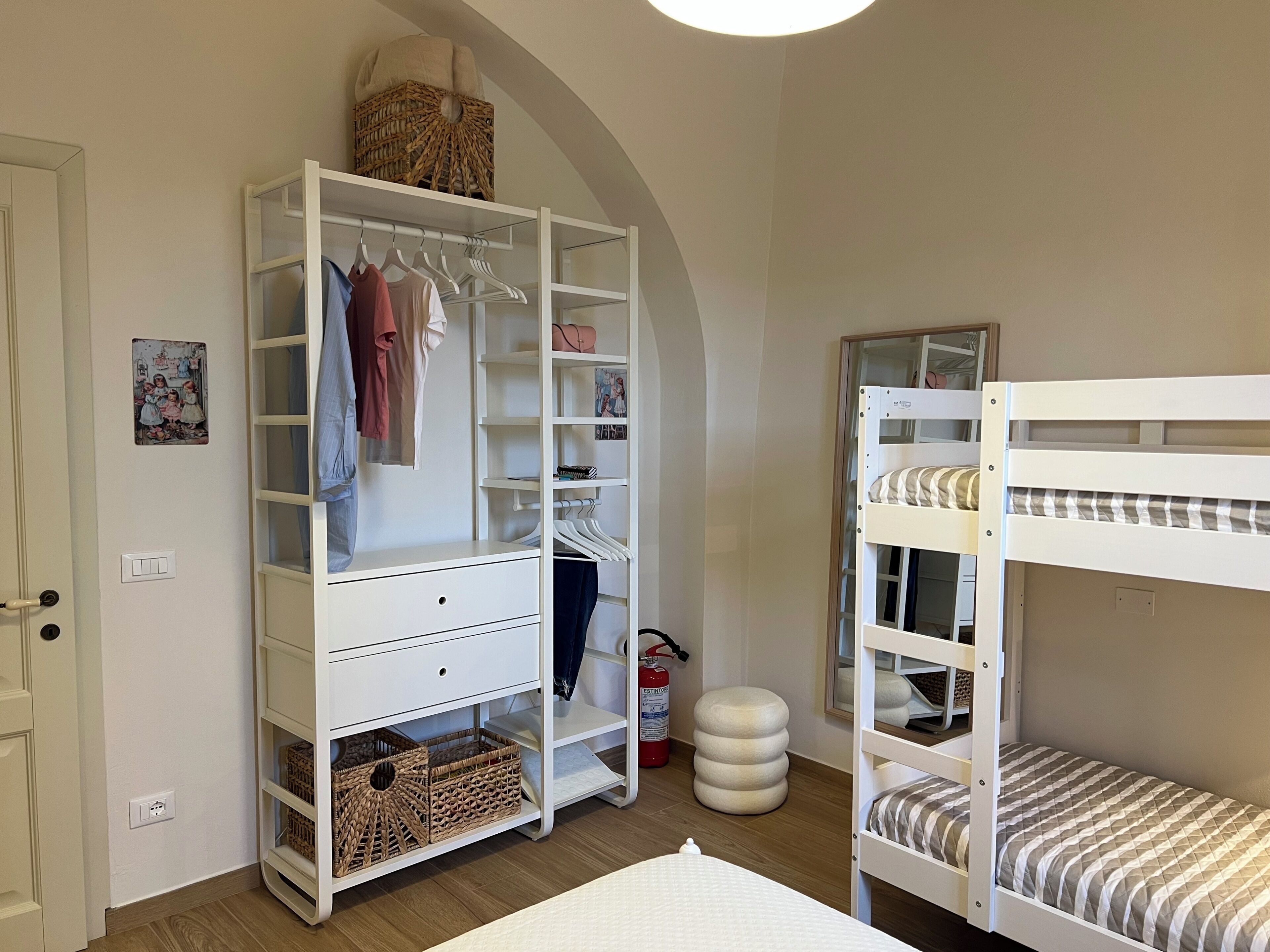 A bedroom with a bunk bed, a white wardrobe with multiple shelves, a small round ottoman, and a wall-mounted fire extinguisher.