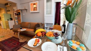 Una sala de estar con sofá, mesa de centro, mesa de comedor con comida y bebidas, y una cocina al fondo.