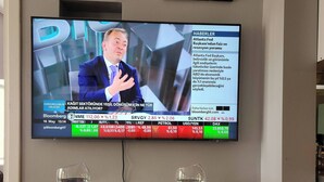 Un televisor montado en la pared mostrando un ticker de noticias de Bloomberg, con dos copas de vino vacías sobre una mesa de vidrio.