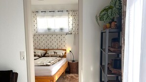 Un dormitori amb un llit, una taula de nit, una làmpada, una finestra amb cortines estampades, una planta i una prestatgeria de fusta.