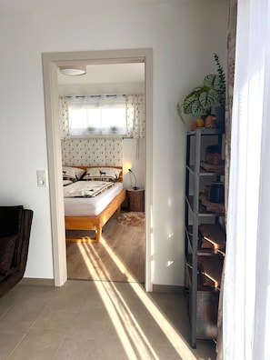 A bedroom with a bed, a bedside table, a lamp, a window with patterned curtains, a plant, and a wooden shelf.