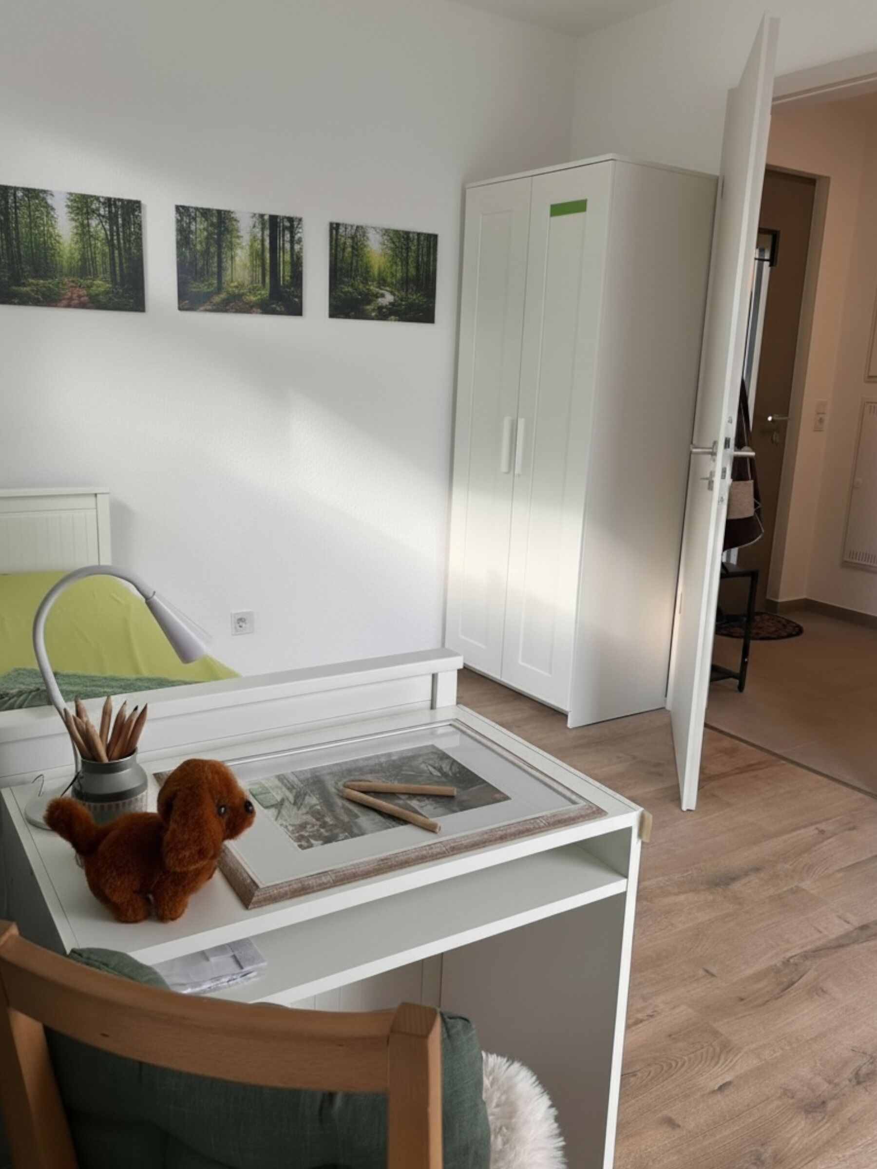 Ett rum med en säng, ett skrivbord med en lampa, en vit garderob och inramade tavlor på väggen.