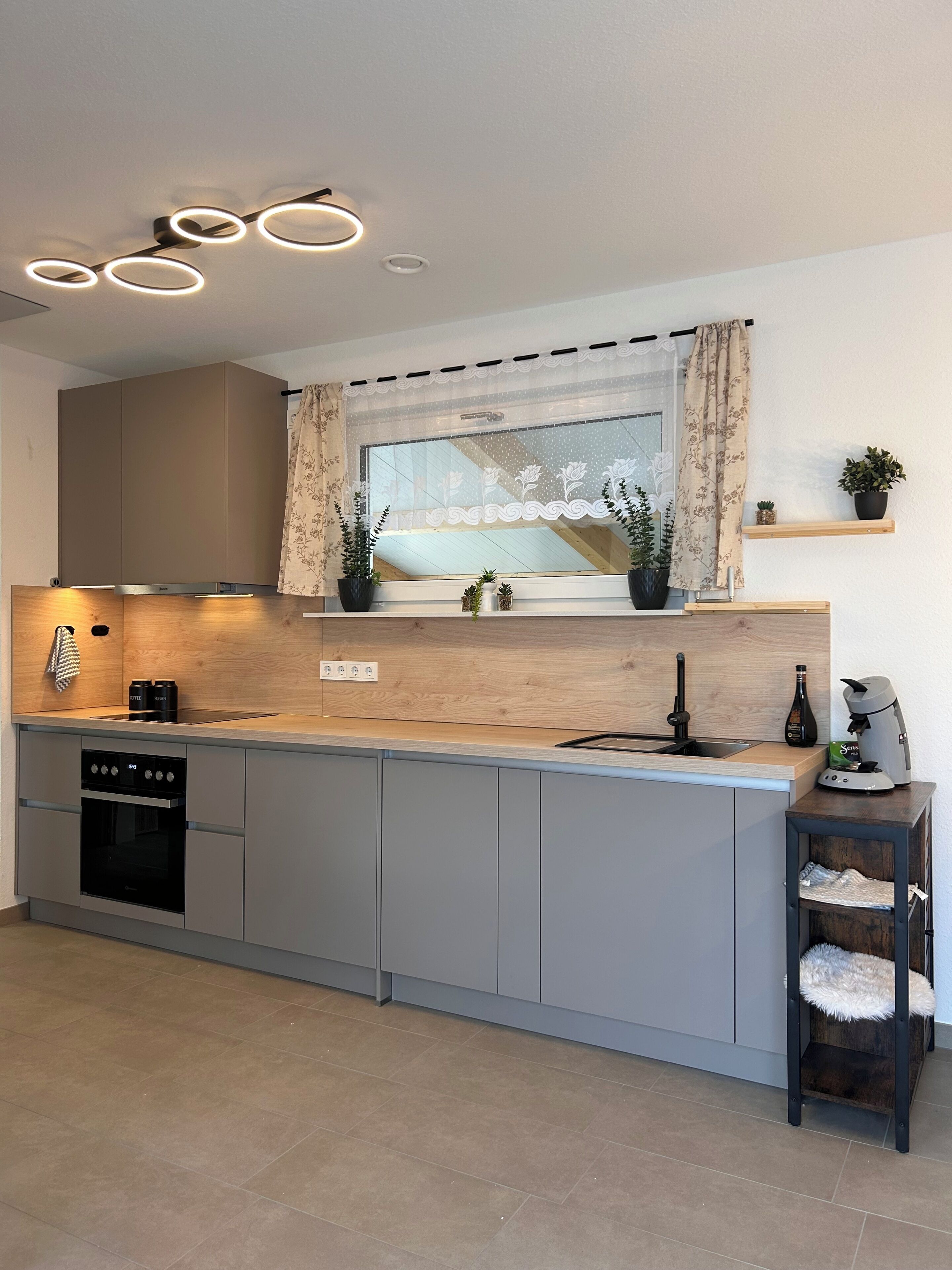 A modern kitchen with a built-in oven, a coffee machine, and a window with curtains.