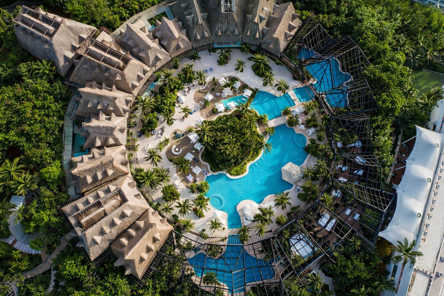 An aerial view of a resort with a central pool, palm trees, and a unique architectural design.