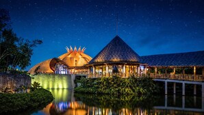 A resort with a thatched-roof building, a dome-shaped structure, and a pavilion with a pointed roof, all reflecting on a calm body of water under a starry night sky. - Grand Mayan Vidanta 2BR Luxury Suite + Pools & Beach (Playa Paraiso)