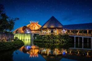Ein Resort mit einem strohgedeckten Gebäude, einer kuppelförmigen Struktur und einem Pavillon mit spitzer Dachform, alles spiegelnd in ruhigem Wasser unter einem sternenübersäten Nachthimmel.