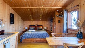 A wooden cabin interior with a bed, a dining table, chairs, a kitchen area, and a window with a view. - Small cottage in the heart of Beaujolais (Rivolet)