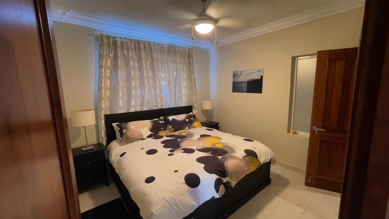 A bedroom with a bed, nightstands, a ceiling fan, a window with curtains, a door, and a wall-mounted picture.
