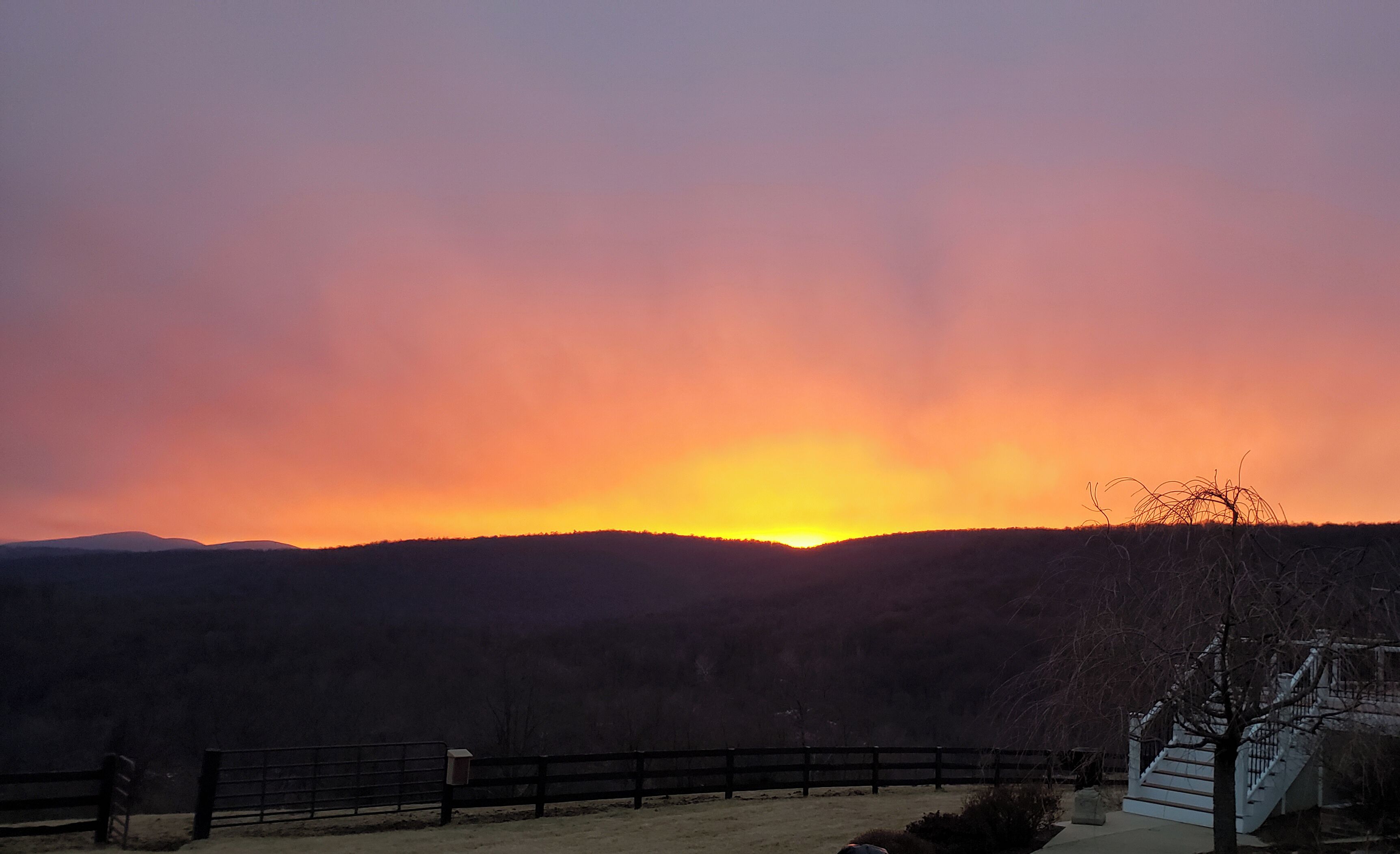 A sunset over a mountainous landscape with a fence and a building in the foreground.