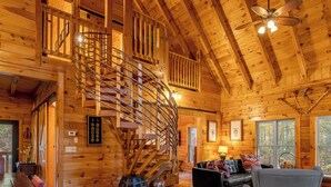 Interior de una cabaña de madera con una escalera de caracol, un comedor, una sala de estar con sofá y chimenea, y ventanas amplias.