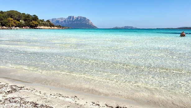 Een strand met helder turquoise water, een zandstrand en een berg op de achtergrond.