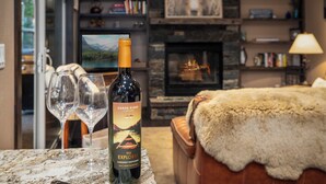 A bottle of wine, two wine glasses, a fireplace, a fur throw, and a bookshelf with books and a bear picture. - Chickadee Cabin - Boutique Stay close to Downtown (Whitefish)