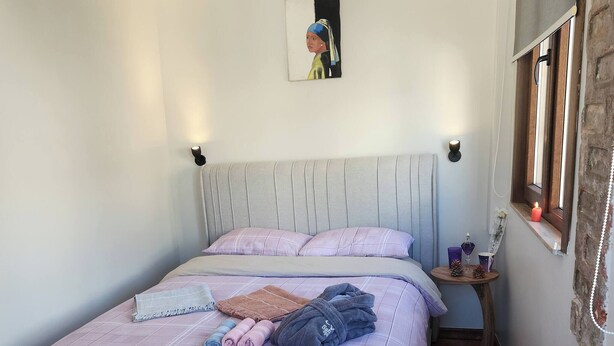 Een bed met een geruit sprei, een nachtkastje met een kaars, en een aan de muur hangend schilderij.