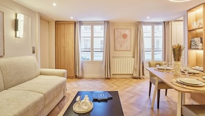Ein modernes Wohnzimmer mit einer Couch, einem Esstisch für zwei Personen und einem großen Fenster mit Vorhängen.