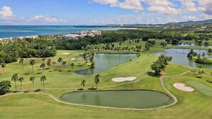 Ein Golfplatz mit mehreren Teichen, Palmen und Blick auf das Meer und die Stadtlandschaft.