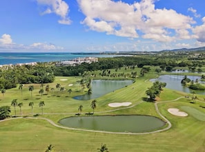 Ein Golfplatz mit mehreren Teichen, Palmen und Blick auf das Meer und die Stadtlandschaft.