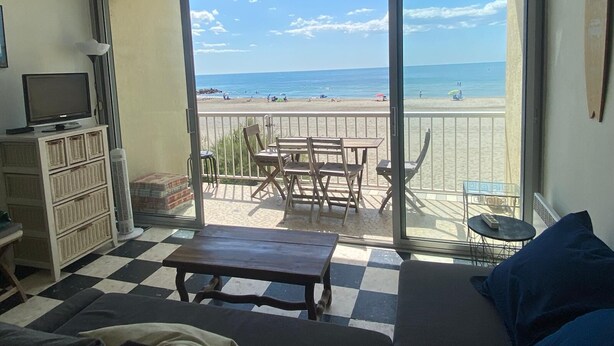 A room with a checkered floor, a TV on a cabinet, a table, chairs, a bed, and a view of the beach through large sliding doors. - Duplex T2 first line on the beach with parking Palavas/Carnon (Palavas-les-Flots)
