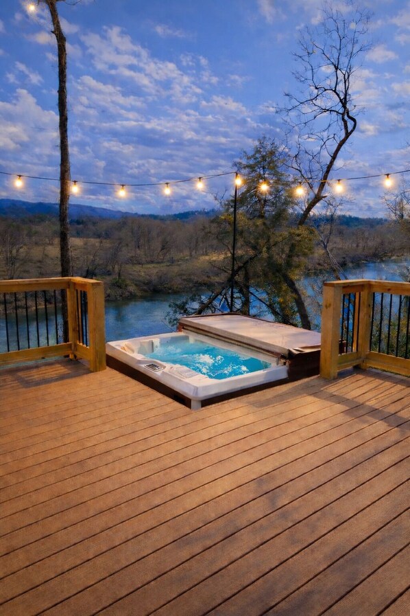 Terraza de madera con jacuzzi e iluminación de luces colgantes, con vista a un cuerpo de agua y árboles.