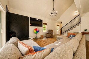 Un salon spacieux avec un grand canapé d’angle, un téléviseur, un lustre et un escalier.